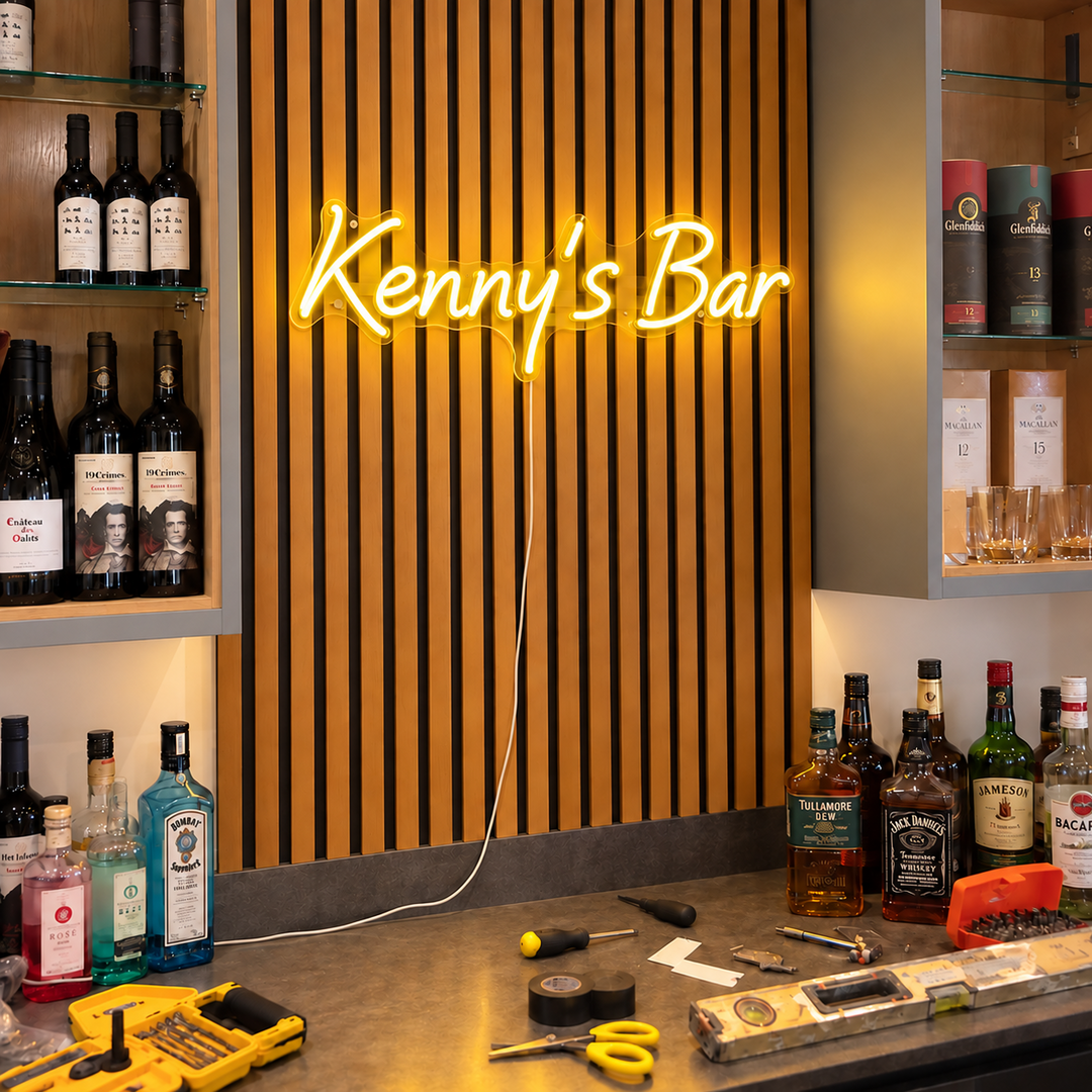 Warm yellow Kenny's Bar neon sign on a wood slat wall between drinks shelves.